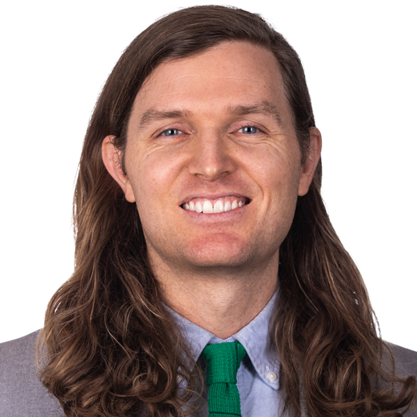 A man in a grey suit, green tie, with long brown hair smiles in front of a white backdrop.