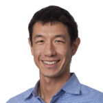 Greg smiles for his headshot in from of a white background.