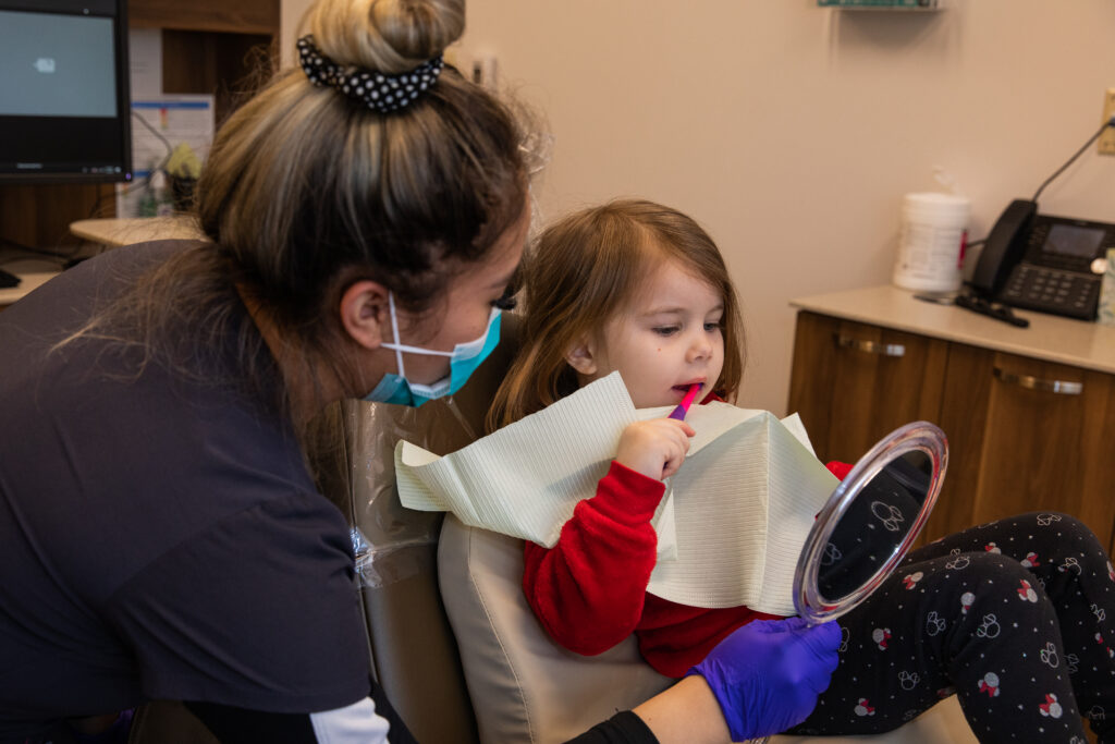 a dental assistant holding up a mirror for a child while they are brushing their teeth