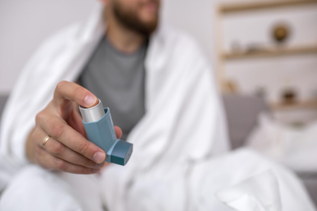 Man bundled in a blanket holding an inhaler, showing breathing difficulties that can worsen in cold weather.