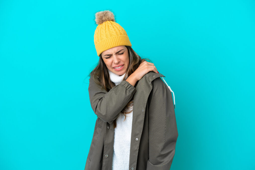 A woman in winter clothing is holding her shoulder in pain from joint stiffness.