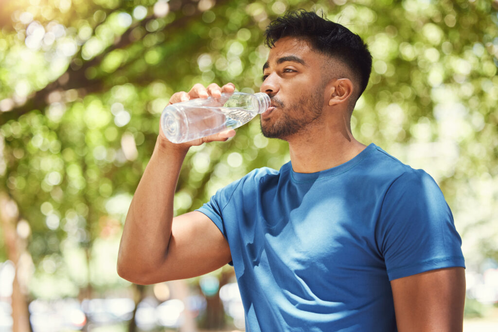 Fitness, man or runner drinking water outdoor for exercise, training or running at a park. Indian male athlete in nature for a workout, run and break to drink from a bottle for health and wellness.